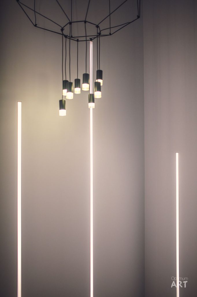 Nowoczesna instalacja oświetleniowa z wieloma lampami wiszącymi na okrągłej konstrukcji sufitowej oraz długimi pionowymi pasami LED na ścianach. Minimalistyczne wnętrze z szarymi ścianami i ciepłym, nastrojowym oświetleniem.