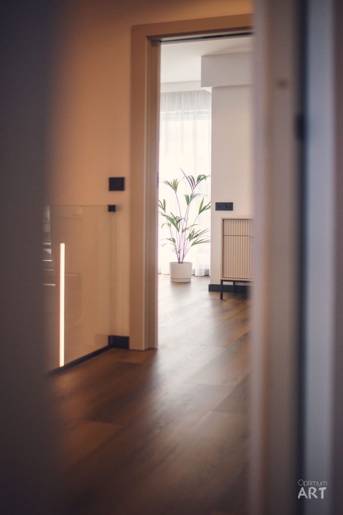 Wnętrze nowoczesnego mieszkania widziane przez otwarte drzwi - jasny korytarz prowadzi do salonu z drewnianą podłogą, gdzie stoi duża roślina doniczkowa i niski stolik. Na ścianie widoczna jest pionowa lampa LED oraz minimalistyczne czarne elementy wyposażenia.