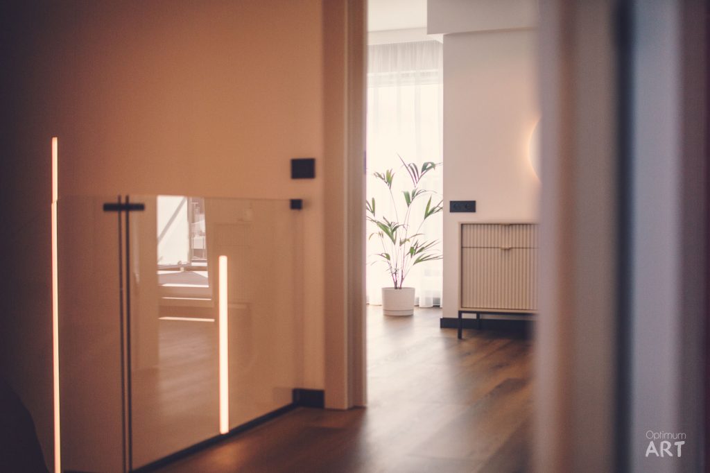 Nowoczesne, minimalistyczne wnętrze z jasnymi drewnianymi elementami i pionowymi lampami LED na ścianach. W tle widoczna roślina w białej doniczce oraz meble o prostych, geometrycznych kształtach na drewnianej podłodze.