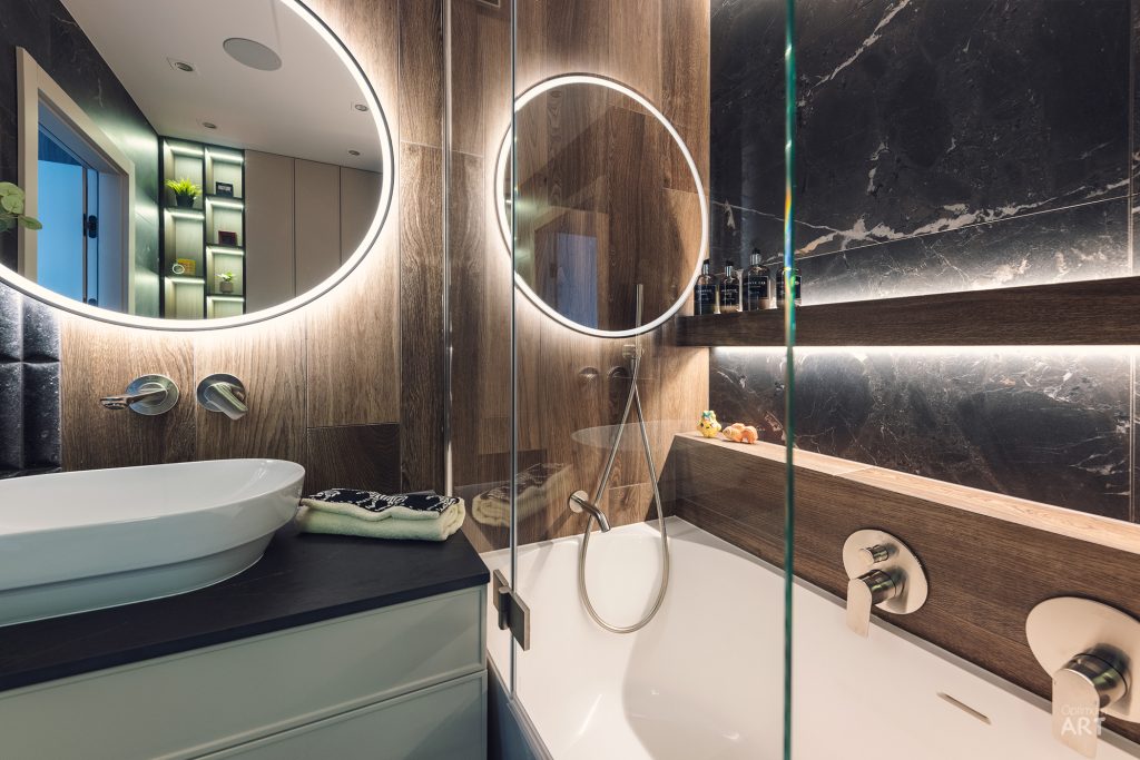 Nowoczesna łazienka z drewnianymi elementami, dwoma okrągłymi lustrami z podświetleniem LED i białą wanną ze szklaną ścianką. Na blacie znajduje się biała umywalka nablatowa, a ściany wykończone są czarnym marmurem i drewnem.