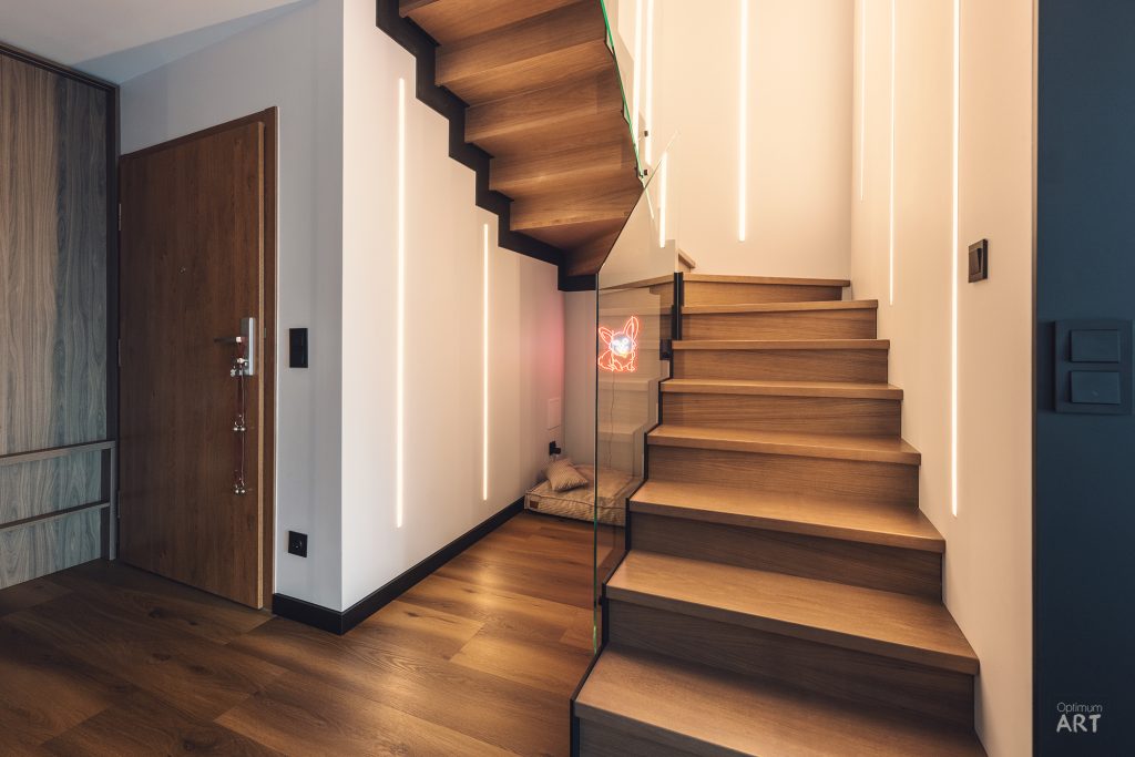 Nowoczesne drewniane schody z podświetleniem LED prowadzą na górę, obok znajdują się drzwi wejściowe z ciemnego drewna. Wnętrze charakteryzuje się minimalistycznym designem z białymi ścianami i ciemnymi akcentami.