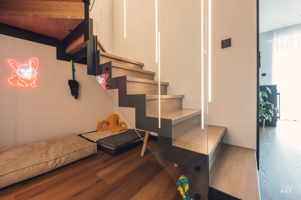 Nowoczesne drewniane schody z czarną stalową konstrukcją prowadzą na wyższe piętro, obok znajduje się drewniana ławka z poduszkami i neonowy znak w kształcie królika na ścianie. Pod schodami widać przestrzeń do przechowywania z zabawkami.