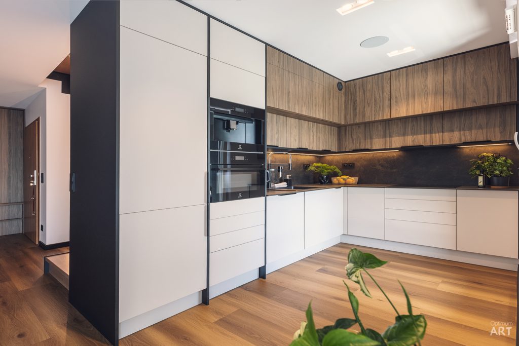 Nowoczesna kuchnia z białymi szafkami dolnymi i drewnianymi górnymi, wyposażona w czarne urządzenia AGD wbudowane w wysoką zabudowę. Pomieszczenie ma drewnianą podłogę, czarny blat z podświetleniem LED i jest ozdobione zielonymi roślinami.