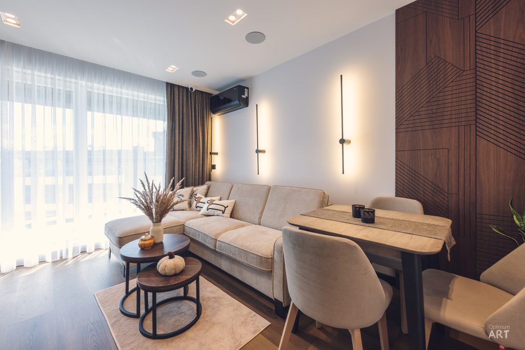 Nowoczesny salon z beżową sofą narożną, okrągłymi stolikami kawowymi i stołem jadalnym z krzesłami. Wnętrze charakteryzuje się drewnianą ścianą z geometrycznym wzorem, dużym oknem z zasłonami i ciepłym oświetleniem.