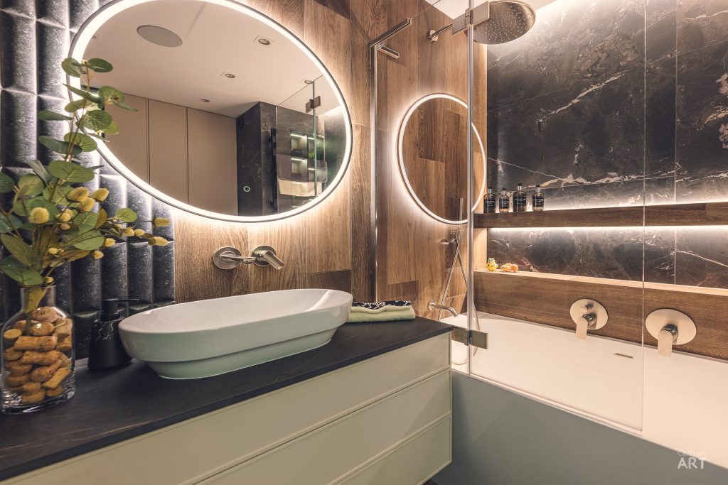 Nowoczesna łazienka z okrągłym lustrem z podświetleniem LED, białą umywalką nablatową i wanną. Wnętrze wykończone jest drewnem i ciemnym marmurem, a po lewej stronie znajduje się roślina doniczkowa.