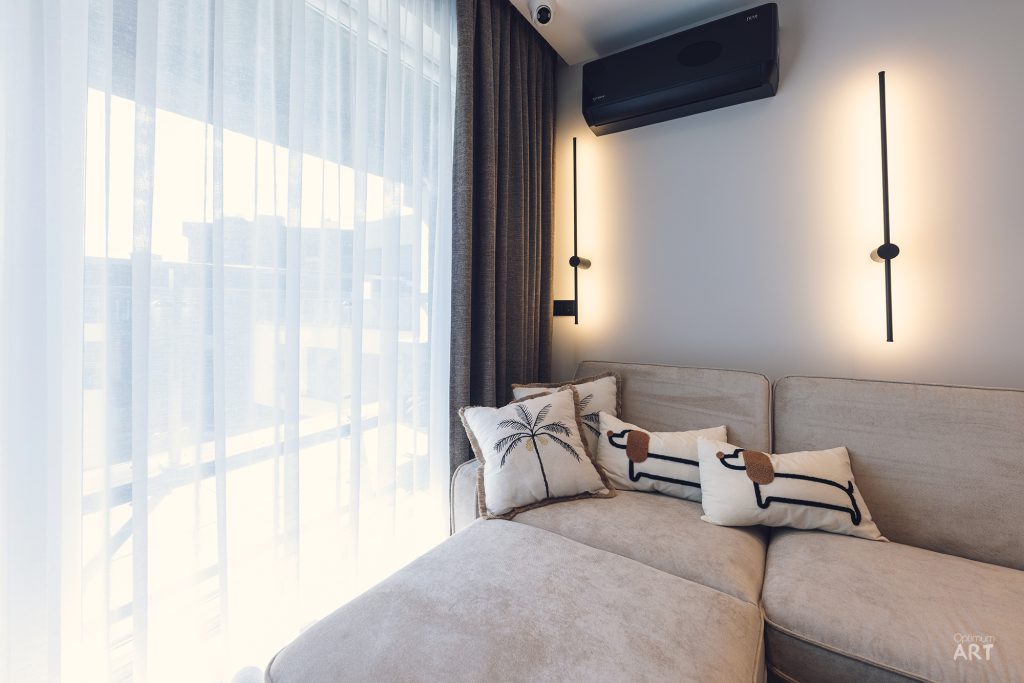 Nowoczesny salon z jasną sofą ustawioną przy oknie z białymi zasłonami i ciemnymi kotarami. Na ścianie wiszą dwie pionowe lampy LED oraz klimatyzator, a na kanapie leżą poduszki z motywem palmy i flamingów.