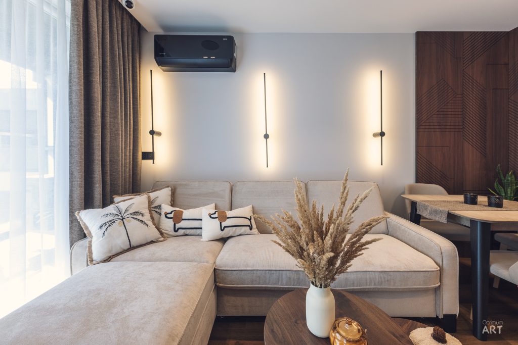 Nowoczesny salon z beżową sofą narożną, na której leżą poduszki dekoracyjne z motywem palmy. Na ścianie wiszą trzy pionowe lampy LED, a na stoliku kawowym stoi wazon z suszoną trawą pampasową.