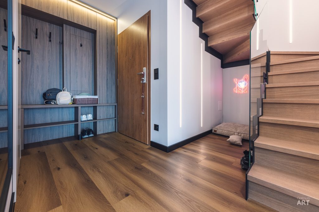 Nowoczesne wnętrze z drewnianymi schodami po prawej stronie i zabudowaną garderobą z wieszakami po lewej. Podłoga wykonana z drewnianych paneli, w tle widoczne drzwi wejściowe i różowy neon na ścianie pod schodami.