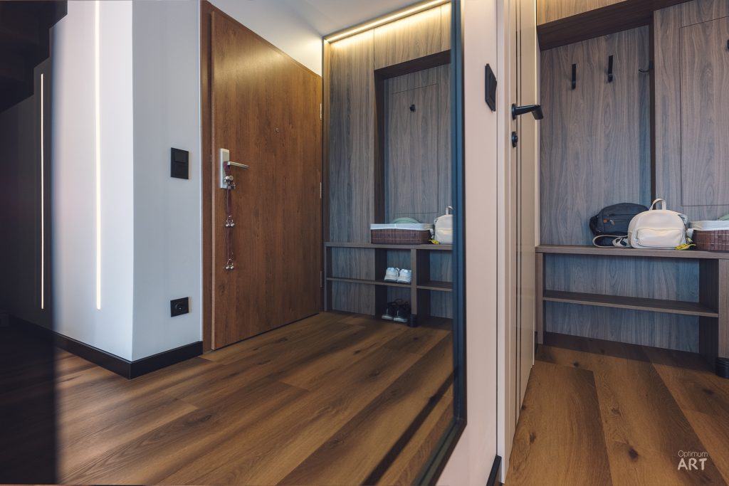 Nowoczesny przedpokój z drewnianą podłogą, białymi ścianami i wbudowanymi szafkami z jasnego drewna. Po prawej stronie znajduje się garderoba z półkami na buty i wieszakami na ubrania, a po lewej drzwi wejściowe z zamkami elektronicznymi.