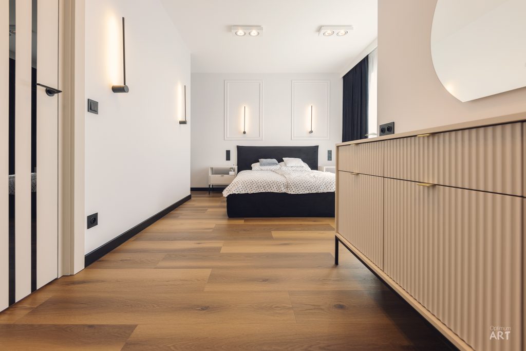 Nowoczesna sypialnia z czarnym łóżkiem na tle białej ściany z dekoracyjnymi panelami i wiszącymi lampami. Po prawej stronie znajduje się drewniana komoda z pionowymi lamelami, a podłoga wykonana jest z jasnego drewna.