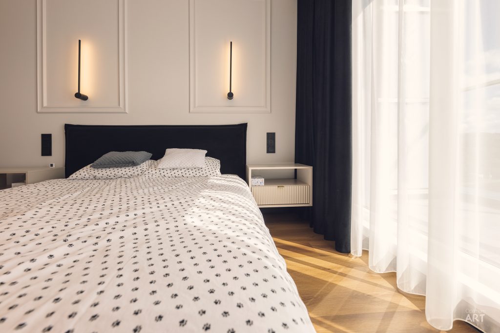 Nowoczesna sypialnia z łóżkiem z czarnym zagłówkiem, białą pościelą w drobny wzór i dwoma minimalistycznymi lampkami ściennymi. Po prawej stronie widoczne są białe zasłony przy oknie, a podłoga wykonana jest z jasnego drewna.