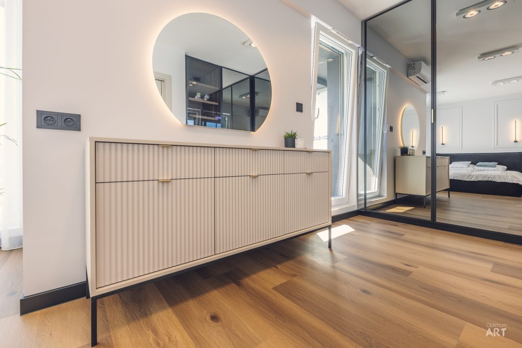 Nowoczesne wnętrze sypialni z jasną drewnianą komodą na tle białej ściany z okrągłym podświetlanym lustrem. Po prawej stronie znajduje się duża szafa z lustrzanymi drzwiami przesuwnymi, a podłoga wykonana jest z jasnego drewna.