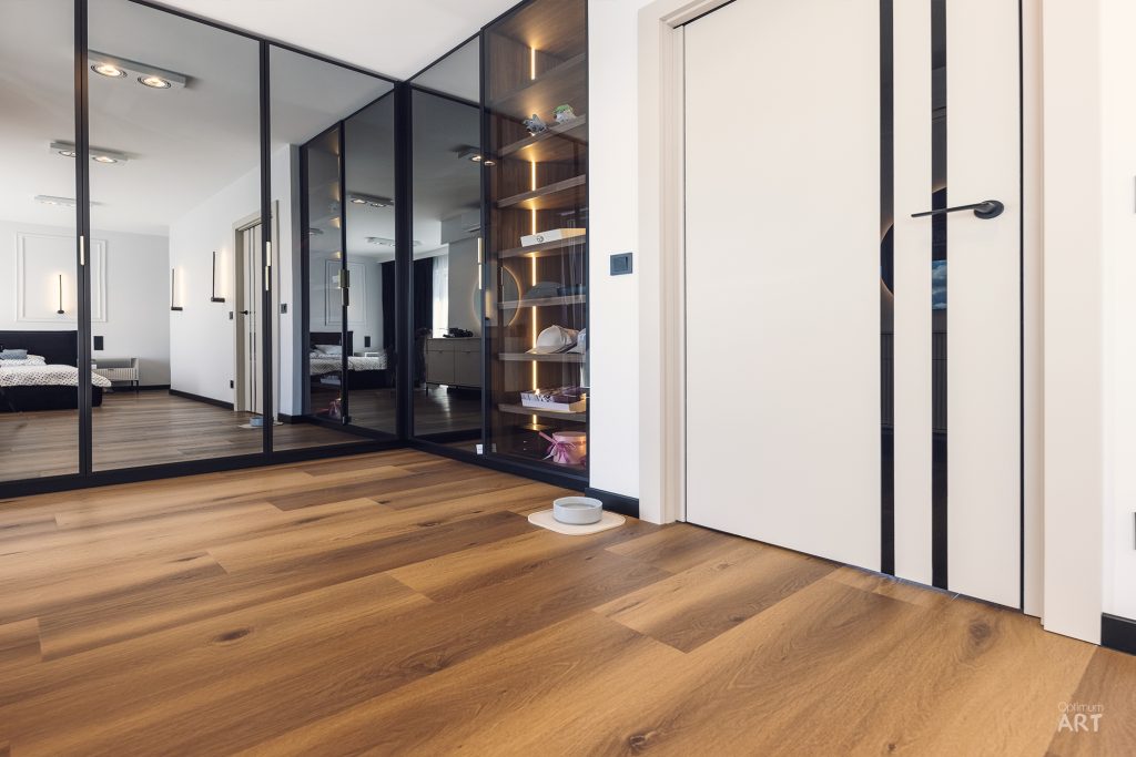 Nowoczesne wnętrze z drewnianą podłogą, dużymi lustrzanymi szafami przesuwanymi z czarnymi ramami po lewej stronie i białymi drzwiami po prawej. W tle widać sypialnię z łóżkiem, a całość utrzymana jest w minimalistycznym stylu z białymi ścianami.