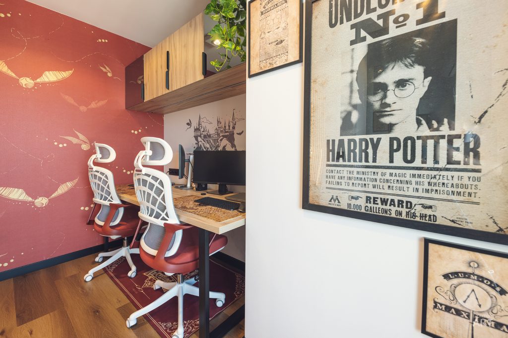 Pokój biurowy z dwoma czerwonymi krzesłami obrotowymi przy drewnianym biurku z komputerem, z czerwoną ścianą ze złotymi motywami z tyłu. Na ścianie wiszą oprawione plakaty w stylu Harry'ego Pottera, w tym plakat poszukiwawczy z wizerunkiem głównego bohatera.
