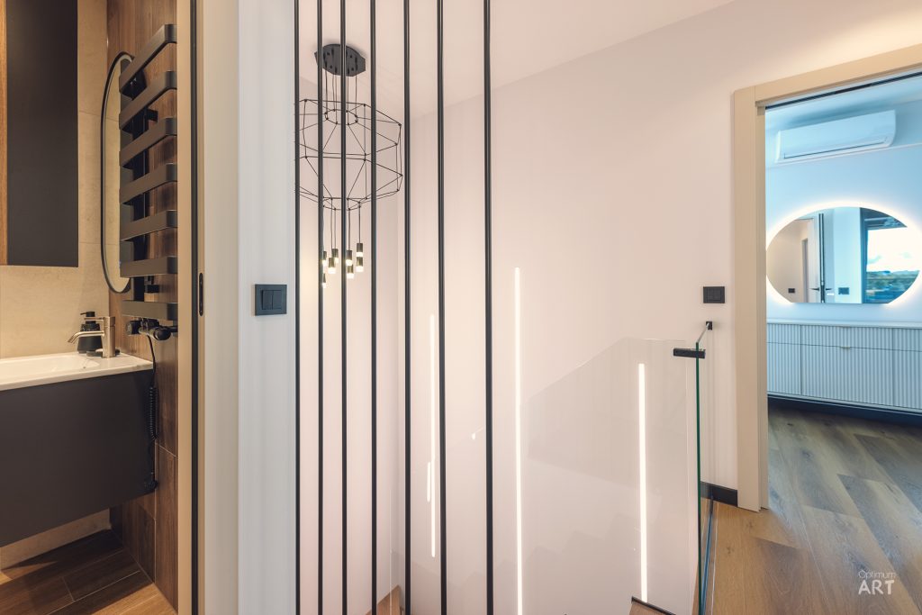 Nowoczesne wnętrze z łazienką po lewej stronie, elegancką przegrodą z pionowymi listewkami w centrum i sypialnią z okrągłym oknem po prawej. Przestrzeń charakteryzuje się jasną kolorystyką, drewnianymi akcentami i minimalistycznym designem.