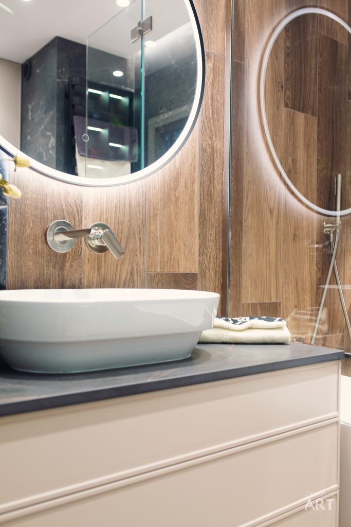 Nowoczesna łazienka z okrągłą umywalką nablatową, drewnianymi elementami wykończeniowymi i podświetlanym lustrem. Na blacie widoczne są złożone ręczniki, a w tle znajduje się kabina prysznicowa ze szklanymi ściankami.