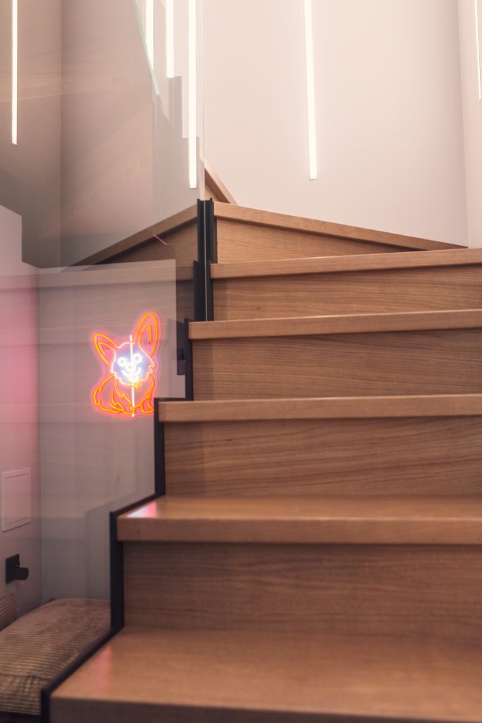 Drewniane schody w nowoczesnym wnętrzu z białymi ścianami i pionowym oświetleniem LED. Na poręczy widoczna jest świetlna dekoracja w kształcie motyla lub kwiatu w kolorach pomarańczowym i różowym.
