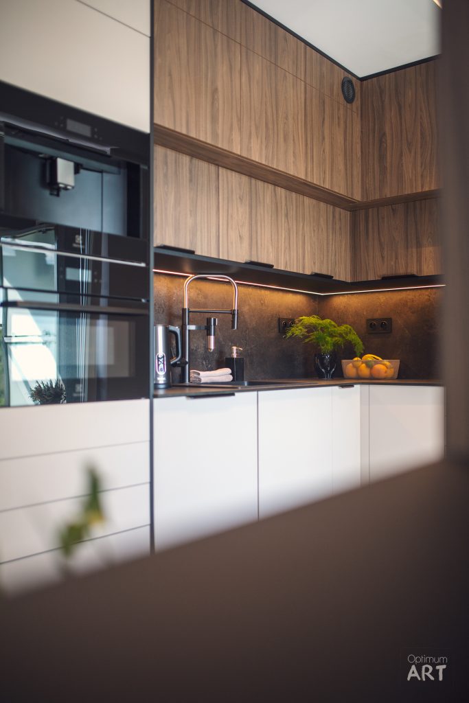 Nowoczesna kuchnia z białymi dolnymi szafkami i górnymi szafkami z drewnianym wykończeniem. Na blacie znajduje się zlewozmywak ze stali nierdzewnej, miska z owocami i wazon z zielonymi roślinami, a po lewej stronie widoczny jest piekarnik.