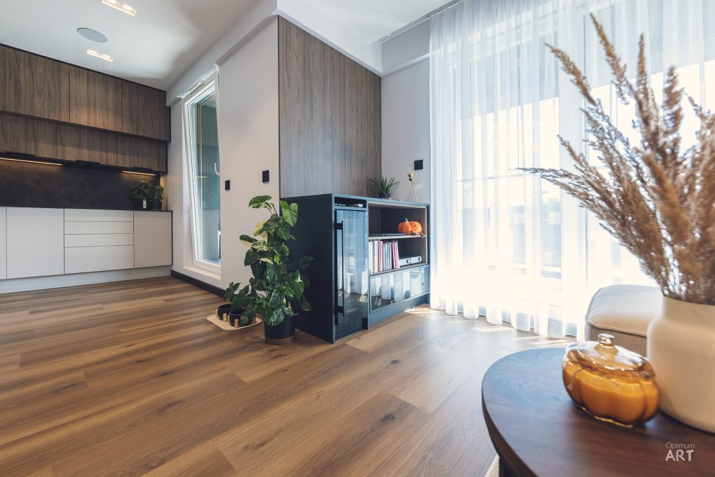 Nowoczesne wnętrze mieszkania z otwartą kuchnią w kolorach drewna i bieli, parkietem na podłodze oraz dużymi oknami z białymi zasłonami. W pomieszczeniu znajdują się rośliny doniczkowe, czarna komoda z książkami oraz okrągły stolik z dekoracyjnymi przedmiotami.