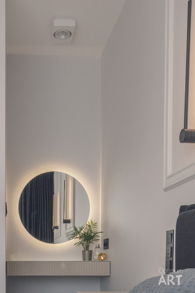 Nowoczesna sypialnia z podświetlanym okrągłym lustrem zamontowanym nad szafką przy ścianie. Pomieszczenie ma białe ściany, a w tle widoczne są ciemne zasłony i część łóżka.