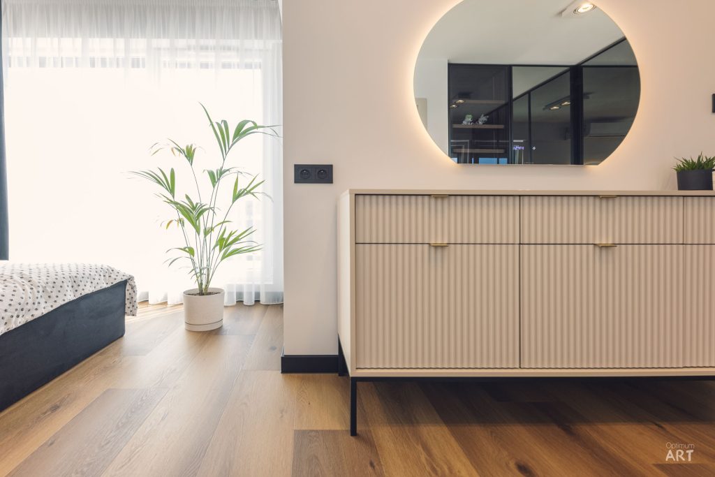 Nowoczesna sypialnia z drewnianą podłogą, łóżkiem z ciemną ramą, jasną komodą z ryflowanymi frontami i okrągłym lustrem z podświetleniem na ścianie. Przy oknie stoi duża roślina doniczkowa, a pomieszczenie jest jasne dzięki naturalnemu światłu.