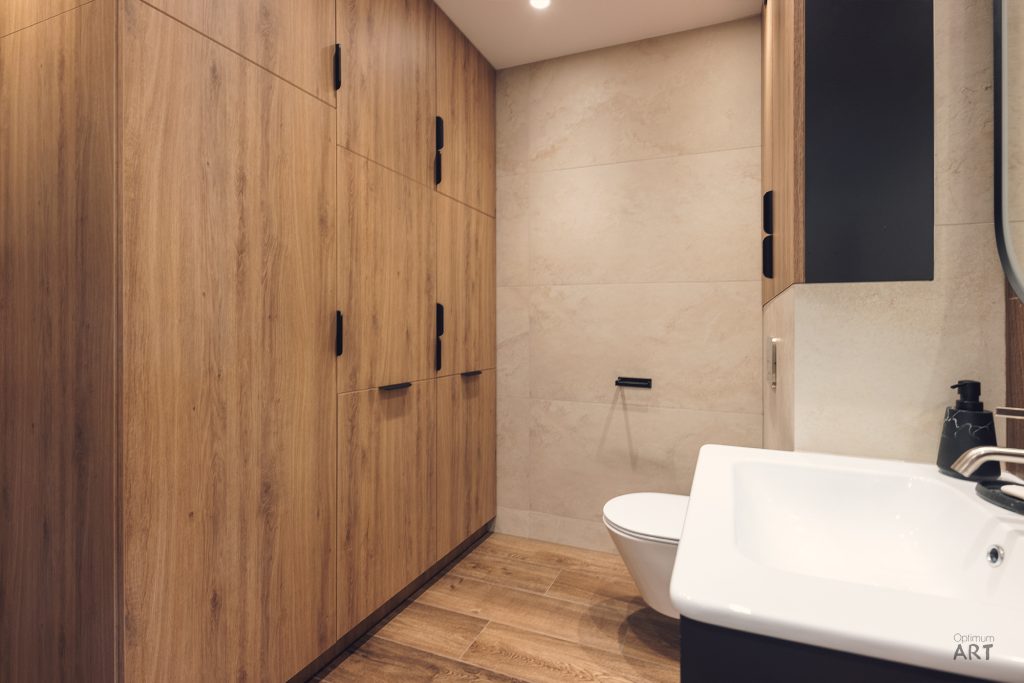 Nowoczesna łazienka z drewnianymi szafkami wzdłuż ściany, białą umywalką nablatową i toaletą w tle. Wnętrze utrzymane w jasnych kolorach z połączeniem drewna, białej ceramiki i beżowych płytek.