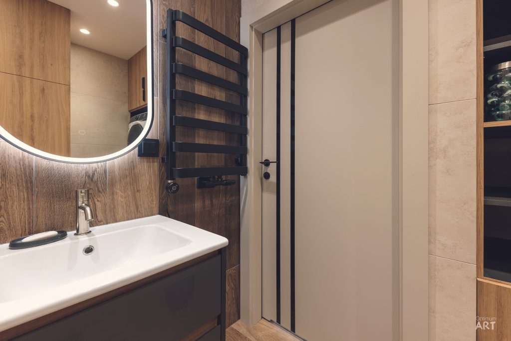 Nowoczesna łazienka z drewnianymi elementami, białą umywalką, okrągłym lustrem z podświetleniem LED i czarnym grzejnikiem ściennym. W tle widoczne są białe drzwi z czarnymi pasami oraz eleganckie wykończenia z drewna i jasnego kamienia.