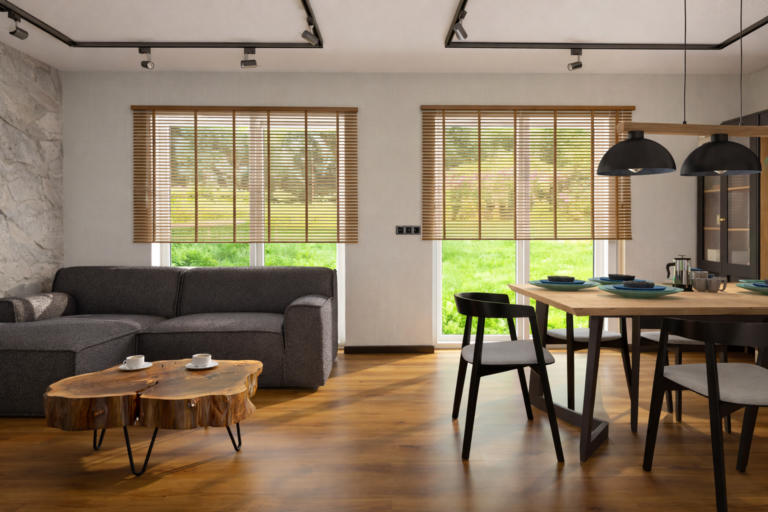 Nowoczesny salon z jadalnią w stylu skandynawskim z szarą sofą, drewnianymi stolikami kawowymi i stołem jadalnym z czarnymi krzesłami. Pomieszczenie ma drewniane podłogi, duże okna z żaluzjami bambusowymi i wiszące czarne lampy nad stołem.