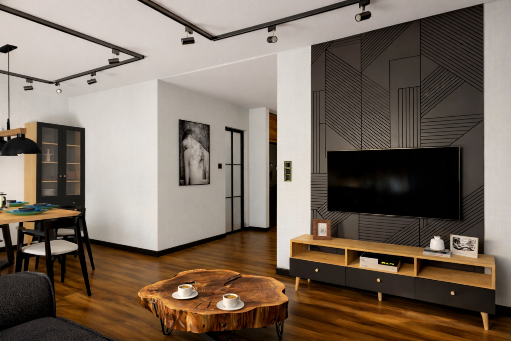 Nowoczesny salon z drewnianą podłogą, na której stoi okrągły stolik z pnia drzewa z filiżankami kawy oraz szara sofa. Na ścianie wisi duży telewizor nad drewnianą szafką RTV, a w tle widać jadalnię ze stołem i krzesłami.