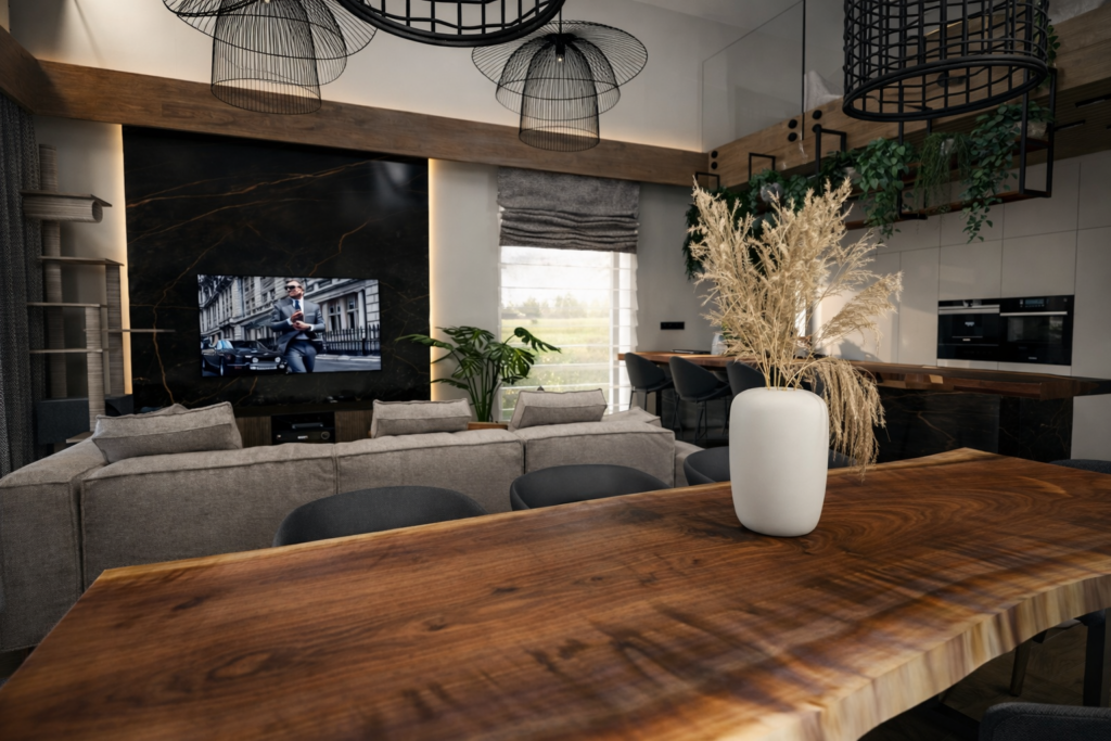 Nowoczesny salon z otwartą kuchnią, gdzie na pierwszym planie znajduje się drewniany stół z białym wazonem pełnym suchych traw. W tle widać szarą sofę zwróconą w stronę telewizora wbudowanego w ciemną ścianę, a nad wszystkim wiszą czarne, ażurowe lampy.