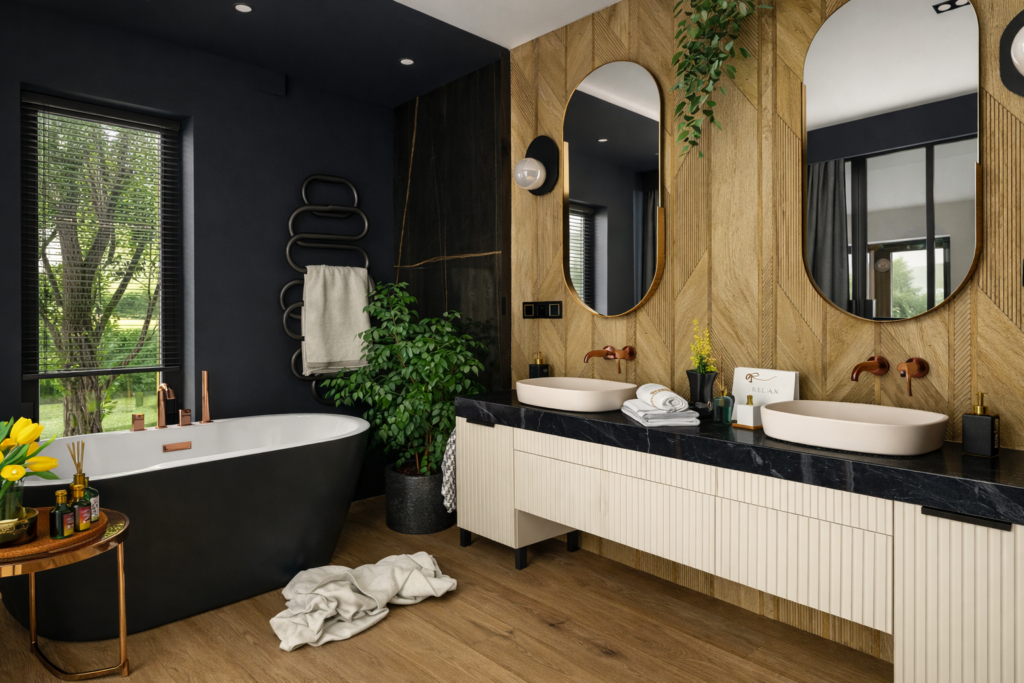 Nowoczesna łazienka z ciemnymi ścianami i drewnianymi akcentami, wyposażona w białą wannę wolnostojącą oraz podwójną umywalkę z okrągłymi lustrami. W pomieszczeniu znajdują się rośliny doniczkowe, drewniany stołek z akcesoriami oraz duże okno z widokiem na zieleń.