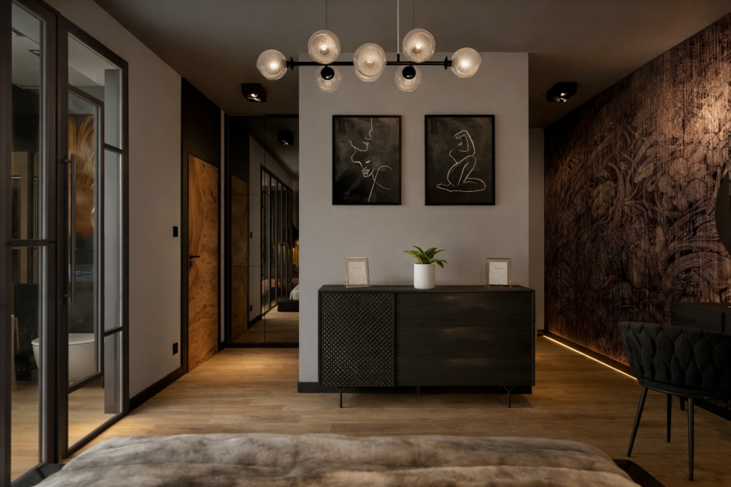 Nowoczesne wnętrze mieszkania z ciemnoszafką pod ścianą, na której wiszą dwa abstrakcyjne obrazy, oraz stylową lampą z okrągłymi kloszami. Po lewej stronie widoczne są przeszklone drzwi, a po prawej ściana z dekoracyjną tapetą i krzesłem.