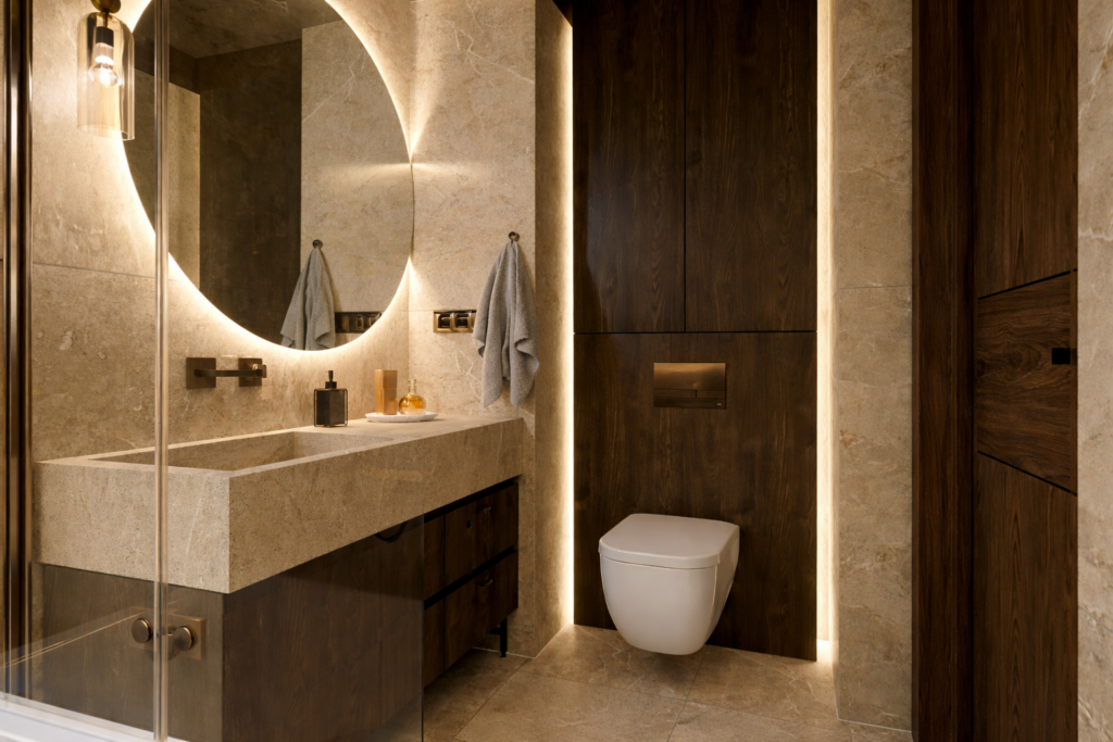 Nowoczesna łazienka z jasnym kamiennym wykończeniem, okrągłym podświetlanym lustrem nad umywalką i wiszącą toaletą w wnęce. Całość utrzymana w ciepłych, beżowo-brązowych tonach z eleganckim oświetleniem LED.